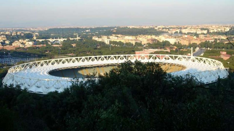 Vista panoramica di uno stadio ovale con struttura esterna bianca a traliccio, circondato da alberi in primo piano. Sullo sfondo si vede un’ampia area urbana con edifici e zone verdi sotto un cielo sereno.