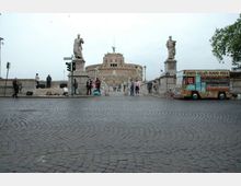 Piazza lastricata in sampietrini con accesso a un ponte, fiancheggiato da due statue su alti piedistalli. Sullo sfondo si vede Castel Sant’Angelo, mentre a destra è parcheggiato un chiosco ambulante per cibo con insegna.