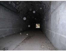 Interno di un tunnel in cemento con volta semicircolare e pareti grezze, illuminato solo da una piccola apertura quadrata in fondo. Il pavimento è coperto di polvere e detriti, con tracce di foglie lungo il passaggio.