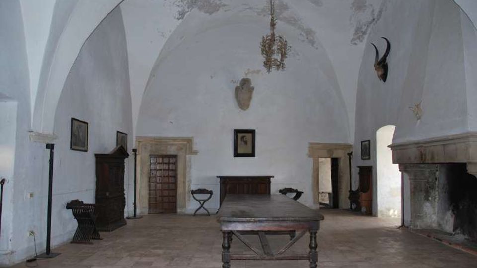 Ampia sala interna con soffitto a volte bianche e pavimento in pietra, arredata con un lungo tavolo centrale in legno e mobili scuri lungo le pareti. Sulla destra si vede un grande camino in pietra e diverse porte e archi che collegano ad altri ambienti.