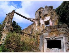 Rovine di un edificio in pietra con muri parzialmente crollati e intonaco consumato, con una trave in legno inclinata tra due pareti. In primo piano si vede un’apertura con cornice in pietra scolpita, circondata da vegetazione e sotto un cielo parzialmente nuvoloso.