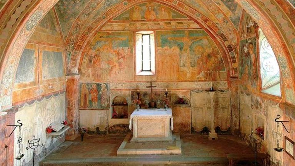 Interno di una piccola cappella con volta a botte e archi ribassati, completamente decorata con affreschi sulle pareti e sul soffitto. In fondo si vede un altare rialzato su gradini, con una finestra centrale stretta e aperture laterali che illuminano l’abside.