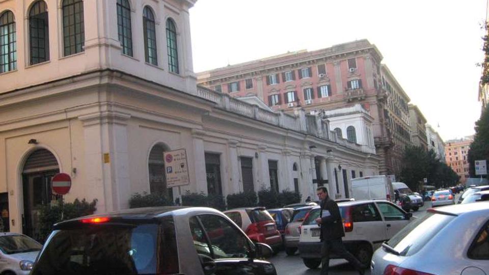 Strada urbana con traffico e auto parcheggiate lungo un edificio storico chiaro con finestre ad arco e facciata lineare. Sullo sfondo si vedono palazzi multipiano e segnaletica stradale in un contesto cittadino.