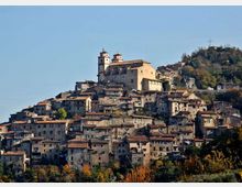 Borgo collinare con case in pietra addossate tra loro e tetti in coppi rossi, disposto a terrazze sul pendio. In cima si vede una grande chiesa con due campanili, con bosco e una croce su un rilievo sullo sfondo.