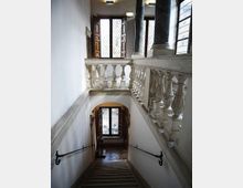 Interno di un edificio storico con scala in pietra che scende verso un pianerottolo, fiancheggiata da una balaustra in marmo e corrimano in ferro. In alto si vedono grandi finestre con griglie e persiane in legno, e colonne scure che sostengono il piano superiore.