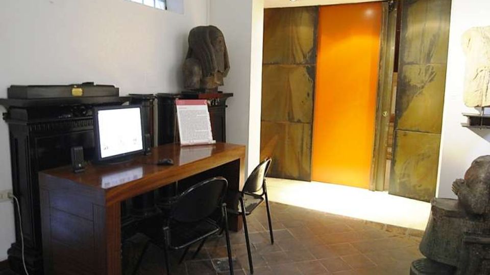 Interno di una sala museale con pavimento in pietra, scrivania in legno con computer e sedie davanti a un mobile scuro. Sul fondo si vede un grande portale con pannello centrale arancione incassato in pareti metalliche, con illuminazione a soffitto e alcune sculture esposte ai lati.