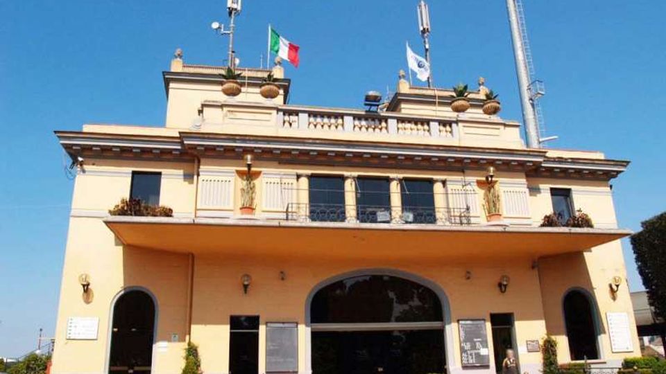 Edificio pubblico a due piani color crema con grande ingresso ad arco centrale, balconata con finestre e terrazza superiore con balaustra. Sul tetto sono presenti antenne e aste con bandiere, con piccolo spazio esterno recintato e panchine davanti alla facciata.