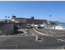 Veduta di un’area portuale con banchine e traghetti attraccati, affiancata da una grande fortificazione in pietra con mura e torri. In primo piano si vede una strada con rotatoria e segnaletica, con il mare e l’orizzonte sullo sfondo.