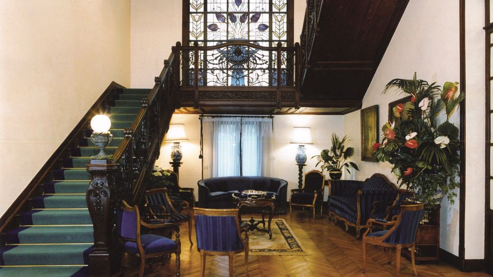 Atrio interno con scala monumentale in legno scuro e moquette verde che porta a un ballatoio con ringhiera decorata, davanti a una grande vetrata istoriata ad arco. Pavimento in parquet a spina di pesce, salottino con divani e poltrone, tappeti e grandi piante ornamentali lungo le pareti.