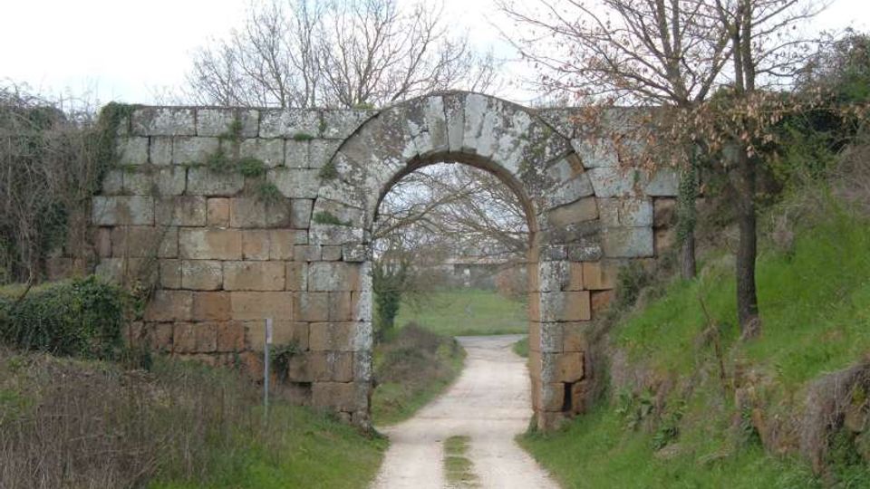 Antico arco in pietra inserito in un basso muro di blocchi squadrati, con tracce di muschio e usura. Un sentiero sterrato passa sotto l’arco e prosegue tra prati e alberi spogli in un paesaggio rurale.