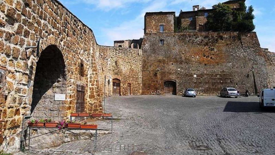 Piazza lastricata in pietra ai piedi di una fortificazione medievale, con alte mura in pietra e un grande arco sul lato sinistro. Sullo sfondo si vede il bastione con edifici in pietra in cima e alcune auto parcheggiate lungo il muro.