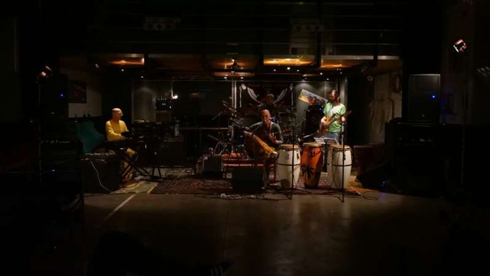 Interno di una sala prove o piccolo studio musicale con palco basso e pavimento lucido, illuminato da luci calde a soffitto. Al centro si vedono batteria, tastiera e strumenti a percussione con microfoni e cavi, affiancati da grandi casse acustiche ai lati.