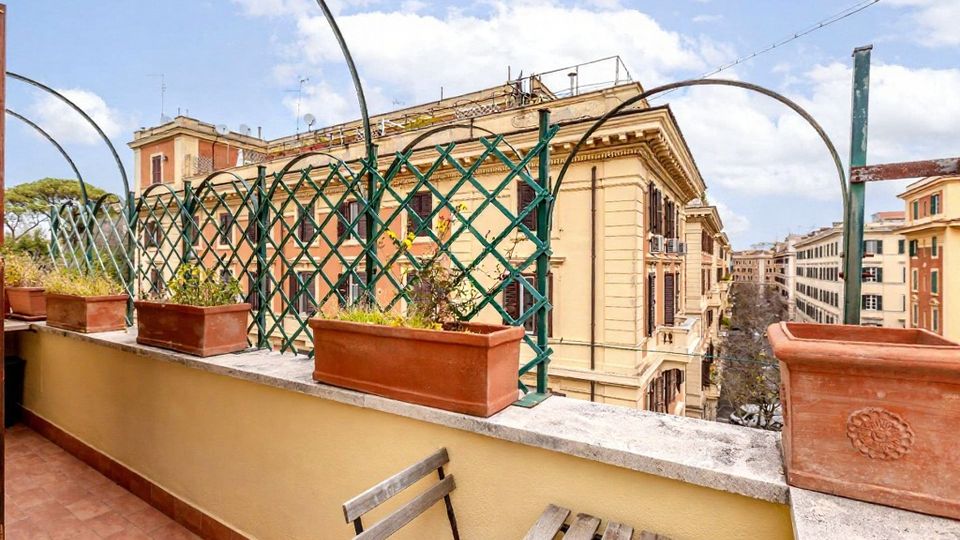Terrazza su tetto con pavimento in cotto, muretto basso e grigliato verde ad arco con fioriere rettangolari. Sullo sfondo si vedono edifici cittadini in stile classico lungo una strada, con cielo parzialmente nuvoloso.