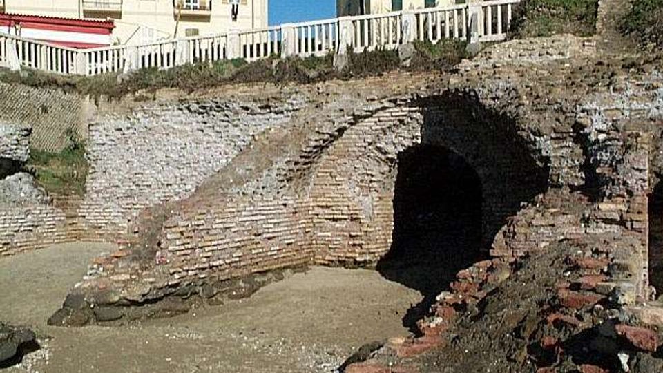 Rovine archeologiche in mattoni con un grande arco che conduce a un passaggio sotterraneo, circondate da muri in pietra. Sullo sfondo si vedono edifici moderni e una ringhiera bianca lungo il bordo superiore del sito.
