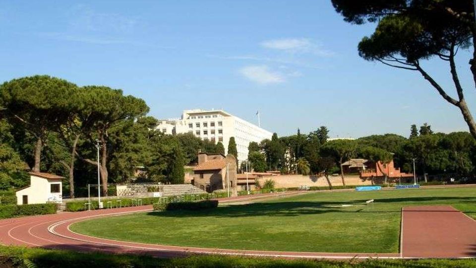 Campo sportivo all’aperto con pista di atletica rossa che circonda un’ampia area erbosa. Sullo sfondo si vedono pini mediterranei, piccoli edifici e una grande struttura bianca multipiano sotto un cielo sereno.