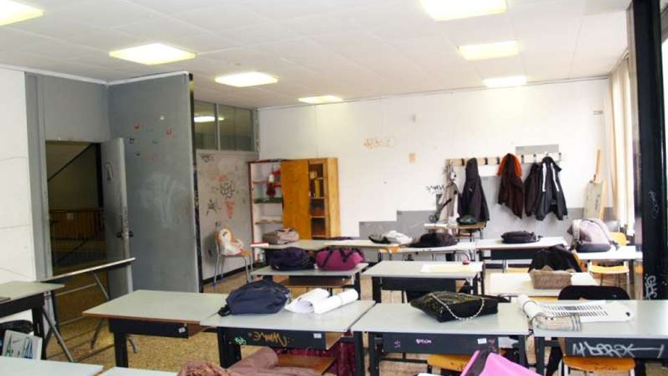 Aula scolastica luminosa con file di banchi e sedie, soffitto a pannelli con luci al neon e pavimento in graniglia. Sul fondo si vedono pareti bianche con appendiabiti e giacche appese, scaffali e una porta aperta verso un corridoio.