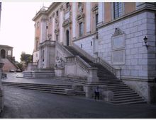Facciata di un grande palazzo storico con pareti in pietra e intonaco, decorazioni scultoree e alte finestre. Un’ampia scalinata monumentale in marmo con balaustre sale dal livello della piazza lastricata fino all’ingresso principale.