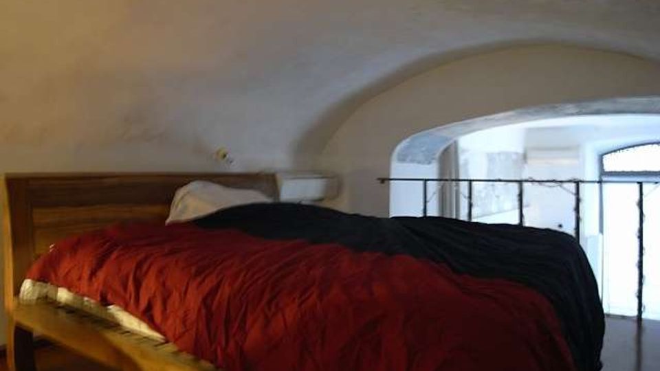 Camera da letto in un soppalco con soffitto a volta intonacato e letto in legno con coperta rossa e nera. Un’apertura ad arco con ringhiera in ferro affaccia su una zona sottostante illuminata da una finestra.