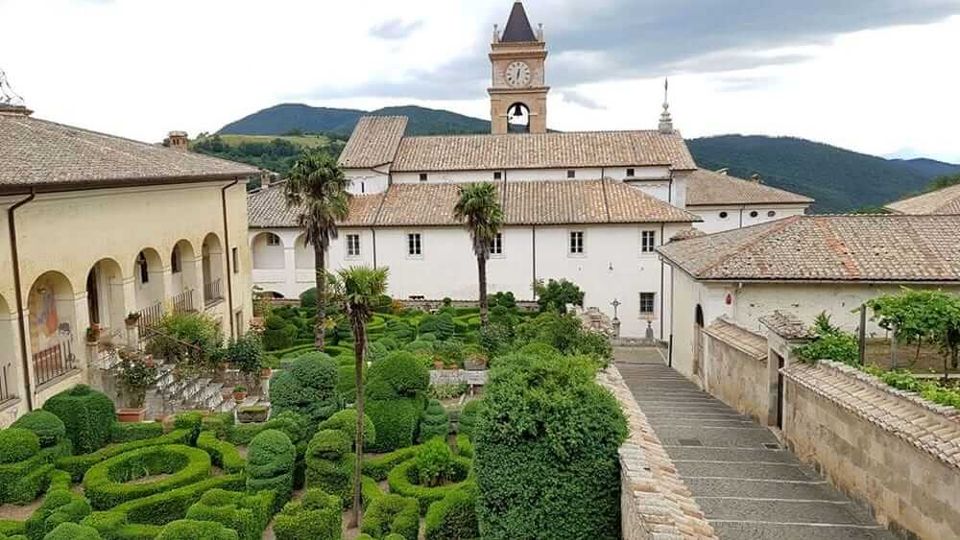 Cortile di complesso monastico con giardino all’italiana di siepi potate a labirinto e alcune palme, circondato da edifici chiari con tetti in coppi. Sullo sfondo si vede una torre dell’orologio e colline verdi sotto un cielo nuvoloso.