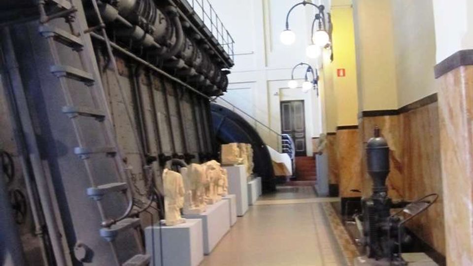 Corridoio interno di un edificio tecnico o museo industriale, con una grande macchina metallica con scaletta e passerella lungo la parete sinistra e modelli esposti su piedistalli. A destra si vedono pareti rivestite in marmo, lampade a globo e una macchina d’epoca, con pavimento a mosaico e una scala sul fondo.