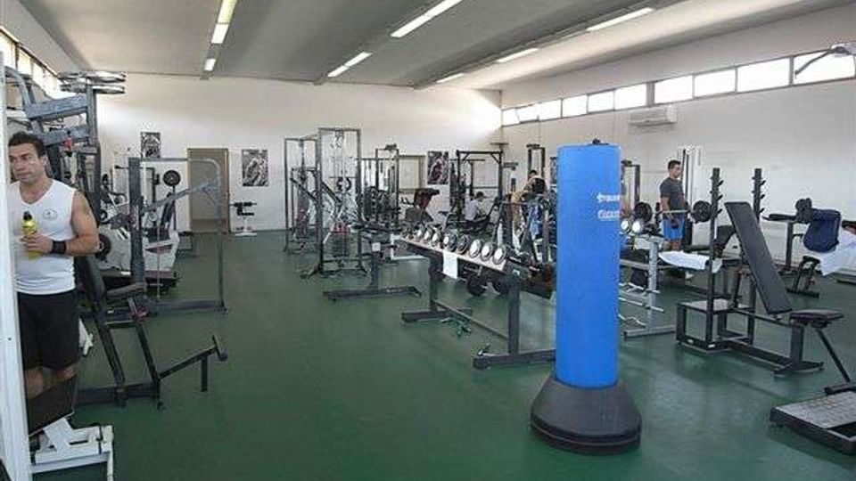 Ampia sala palestra con pavimento verde e soffitto alto illuminato da lunghe luci al neon, con grandi finestre lungo la parete destra. Sono presenti varie attrezzature da allenamento come panche, rastrelliere, macchine per pesi e un sacco da boxe blu al centro.