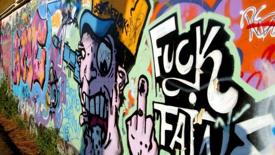Muro esterno lungo una strada ricoperto da graffiti multicolori e street art, con grandi scritte e un personaggio illustrato. Il muro corre in prospettiva con vegetazione bassa alla base e superficie in cemento visibile.