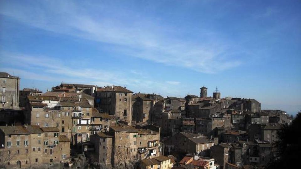 Veduta panoramica di un borgo collinare con edifici in pietra addossati tra loro e tetti in coppi, con alcune torri e campanili che emergono sopra le case. Sullo sfondo si vede un ampio cielo azzurro e l’orizzonte in lontananza.