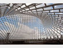 Ampia struttura architettonica moderna con grande copertura a reticolo in acciaio che forma una volta curva, aperta sui lati e verso il cielo. In basso si vedono gradinate e una base in cemento, come un’area eventi o anfiteatro in costruzione.