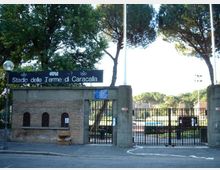 Ingresso dello Stadio delle Terme di Caracalla con cancello in ferro e muri in mattoni e cemento, sormontato da un’insegna con il nome dello stadio. Sullo sfondo si vedono alberi alti e parte dell’area sportiva interna.
