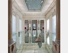Ampia sala interna in stile neoclassico con alte colonne, grandi finestre con tende chiare e soffitto a cassettoni con lucernario centrale. Pavimento lucido in marmo venato con piccole sculture bianche disposte nello spazio e riflessi sull’acqua o su una superficie molto brillante.