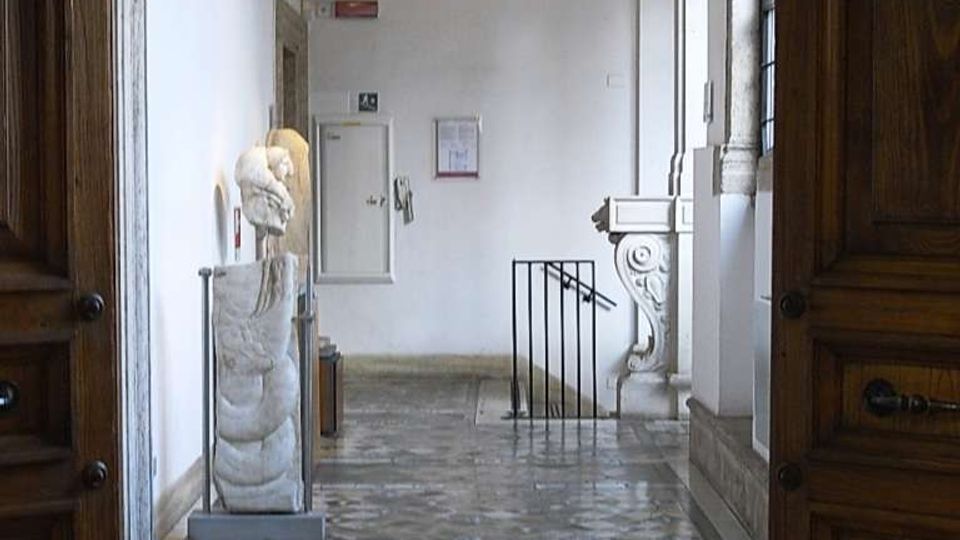 Interno di un edificio storico visto attraverso un grande portone in legno, con pavimento a mosaico geometrico e pareti bianche. Lungo il corridoio si notano una scultura su piedistallo, colonne e finestre alte, e una ringhiera che delimita una scala.