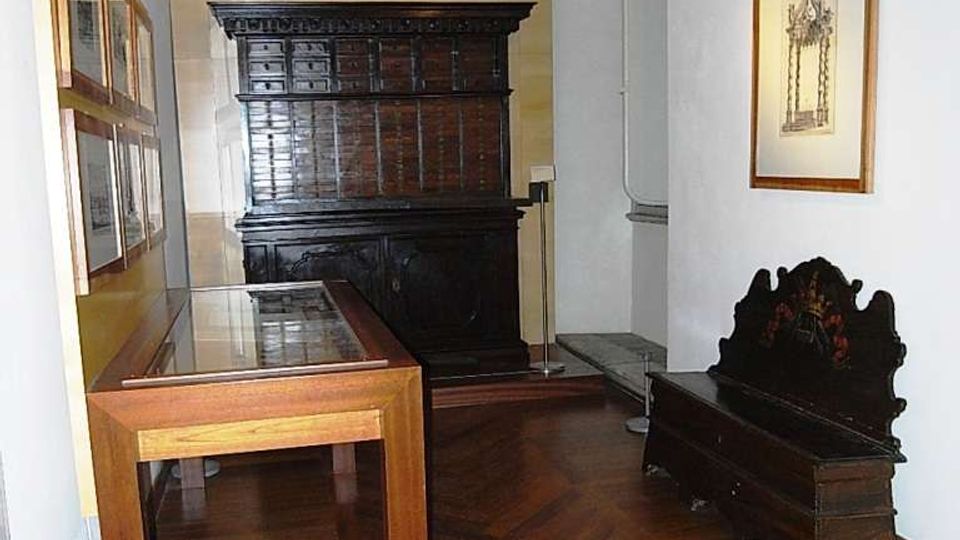 Interno di una piccola sala museale con pareti bianche e pavimento in parquet a spina di pesce, arredato con una grande credenza scura, una panca antica e una vetrina espositiva in legno e vetro. Sulle pareti sono appesi quadri incorniciati e l’illuminazione è a soffitto alto.