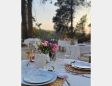 Tavolo apparecchiato all’aperto con tovaglia bianca, piatti, bicchieri e posate su sottopiatti intrecciati, decorato con un vaso di fiori e una lanterna. Sullo sfondo si vedono altri tavoli disposti in un’area verde con alberi al tramonto.