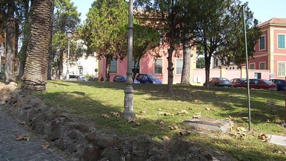 Piccolo parco urbano con prato in lieve pendenza, alberi alti e un lampione centrale, bordato da un muretto di pietra e da una pavimentazione in ciottoli. Sullo sfondo si vedono edifici con facciate rosa e bianche e alcune auto parcheggiate lungo la strada.