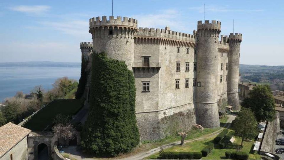 Veduta esterna di un grande castello in pietra con torri cilindriche merlate e mura alte, affacciato su una collina con giardini e un vialetto d’accesso. Sullo sfondo si vede un lago e un paesaggio collinare sotto un cielo sereno.