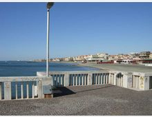 Belvedere sul lungomare con pavimentazione in pietra e balaustra in cemento, con un lampione al centro in primo piano. Sullo sfondo si vede la costa con spiaggia, mare calmo e una fila di edifici lungo la riva sotto un cielo sereno.
