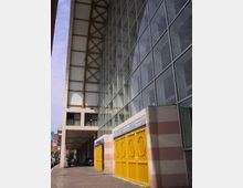 Facciata di un grande edificio moderno con parete vetrata a griglia e ampia pensilina metallica sostenuta da travi. Al piano strada si vedono ingressi con cancelli gialli e un porticato lungo il marciapiede in ambiente urbano.