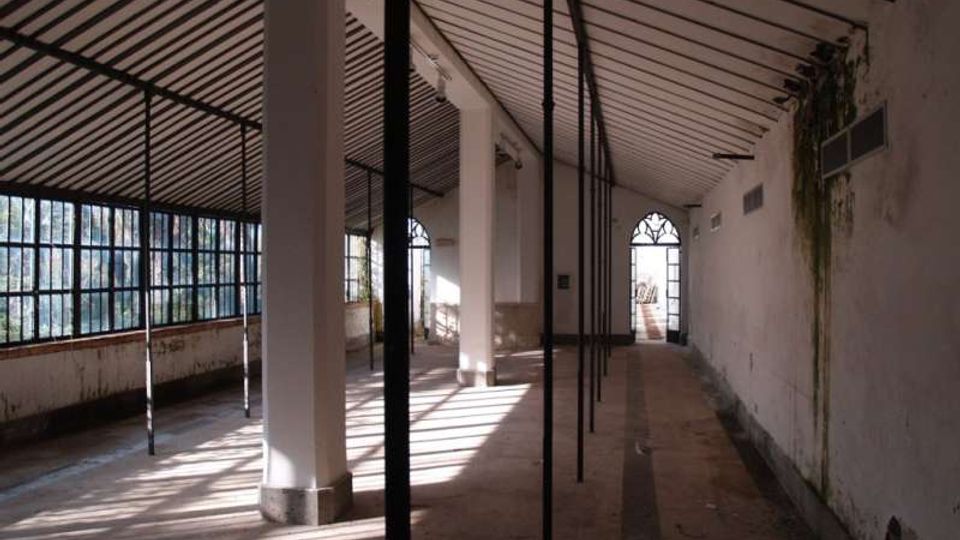 Interno di un lungo spazio industriale dismesso con soffitto a falde e travi a vista, pilastri bianchi e sottili montanti metallici in fila. Lungo il lato sinistro ci sono ampie finestre a griglia che lasciano entrare luce radente, mentre sul fondo si vede una porta ad arco.