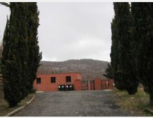 Ingresso asfaltato fiancheggiato da alti cipressi che conduce a un edificio basso in mattoni rossi con finestre rettangolari e un cancello metallico. Sullo sfondo si vedono colline boscate sotto un cielo coperto.