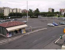 Ampio parcheggio asfaltato con strisce di sosta e un’edicola a un piano con tetto in tegole rosse in primo piano. Sullo sfondo si vedono alberi, strade di raccordo e palazzi residenziali e uffici.