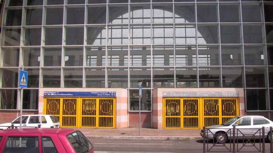 Facciata di un grande edificio pubblico con ampia vetrata a griglia che copre un alto arco centrale. Alla base ci sono due ingressi con cancelli gialli, insegne sopra le porte e marciapiede su strada con auto parcheggiate e segnaletica pedonale.