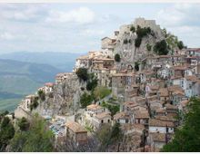 Borgo storico arroccato su uno sperone roccioso, con case in pietra dai tetti in coppi addossate lungo il pendio e una fortificazione in cima. Sullo sfondo si vedono colline e montagne, con una strada e alcune auto ai piedi della rupe.