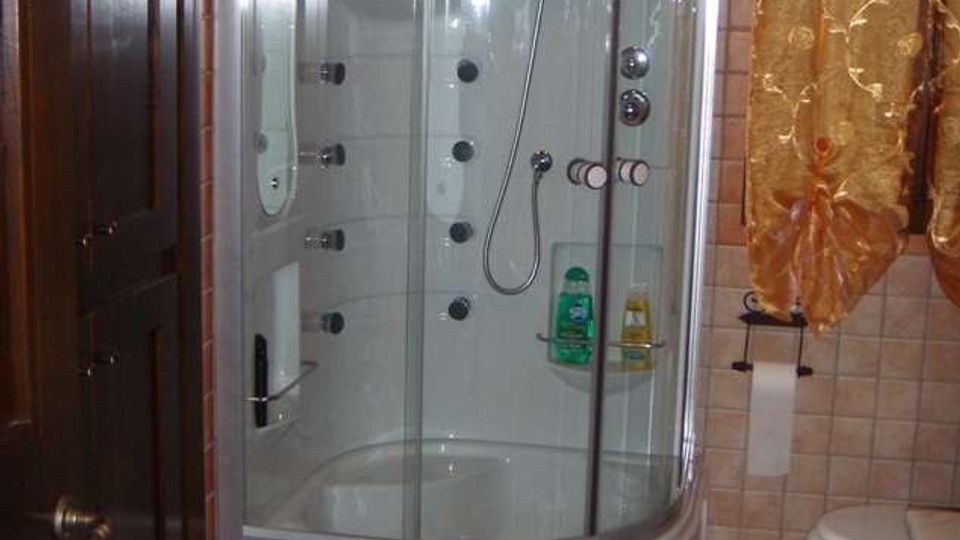 Bagno con cabina doccia angolare in vetro curvo e base bianca, dotata di soffioni e getti idromassaggio interni. Pareti e pavimento rivestiti in piastrelle color beige, con porta in legno scuro e sanitari visibili sul lato destro.