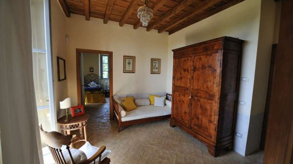 Interno di una camera in stile rustico con soffitto in legno a travi e lampadario, pavimento in cotto e pareti chiare. Arredamento con grande armadio in legno, divanetto con cuscini e tavolino vicino a una finestra, con porta aperta verso una stanza adiacente.