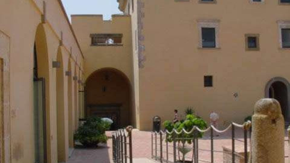 Cortile interno con pavimentazione in mattoni e passaggio centrale delimitato da una ringhiera metallica, affiancato da un edificio storico giallo con arcate e finestre. In primo piano si vede una colonna o statua in pietra e sullo sfondo un portico con grande arco che conduce a un’area ombreggiata.