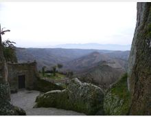 Veduta panoramica di una valle collinare con rilievi e creste in lontananza, osservata da un belvedere in pietra. In primo piano si vedono rocce ricoperte di muschio e un piccolo edificio in muratura con terrazza e muretto.