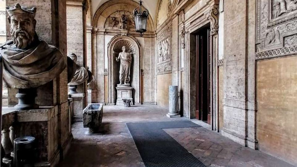 Porticato storico in pietra con soffitto a volte e pavimento in mattoni, fiancheggiato da busti e statue in nicchie. Sulle pareti sono visibili rilievi decorativi e un grande ingresso ad arco, con lanterna sospesa al centro del corridoio.