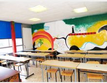 Aula scolastica con file di banchi e sedie in legno su pavimento in graniglia, illuminata da plafoniere a soffitto e con grandi finestre schermate da tende blu. La parete di fondo è decorata con un ampio murale colorato che raffigura forme astratte, un arcobaleno e un faro su uno sfondo giallo e verde.