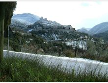 Vista panoramica di un borgo in pietra arroccato su una collina, con edifici compatti e una torre centrale, circondato da pendii e uliveti leggermente innevati. In primo piano si vede una strada asfaltata con erba alta ai bordi e segnaletica stradale, sullo sfondo colline e montagne.