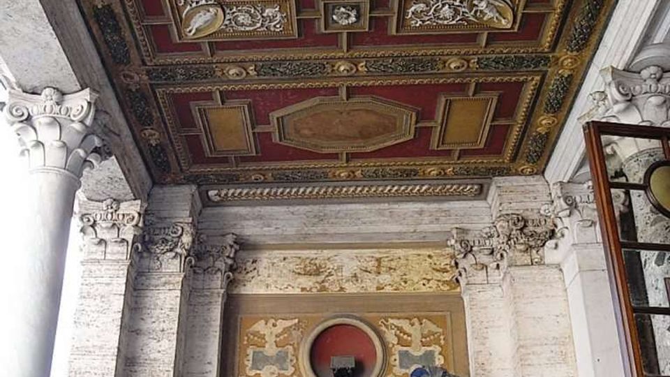 Soffitto a cassettoni riccamente decorato in rosso e oro sopra un portico in pietra con colonne e capitelli scolpiti. In basso si vede un ingresso con portale in pietra e doppia porta in legno, sormontato da un fregio ornamentale.